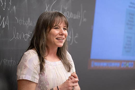 Photo of a professor smiling at the front of a classroom, instructing students off-screen with a chalkboard behind her