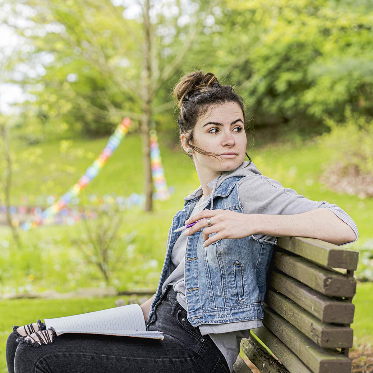 查塔姆学院一名女学生手里拿着铅笔的照片, 她坐在阴凉校园外的长凳上，膝上放着一本打开的笔记本