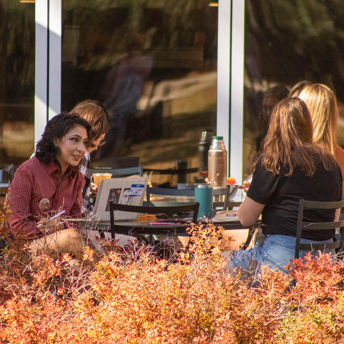 一群学生坐在咖啡厅外的桌椅上, 前景是秋天的树叶