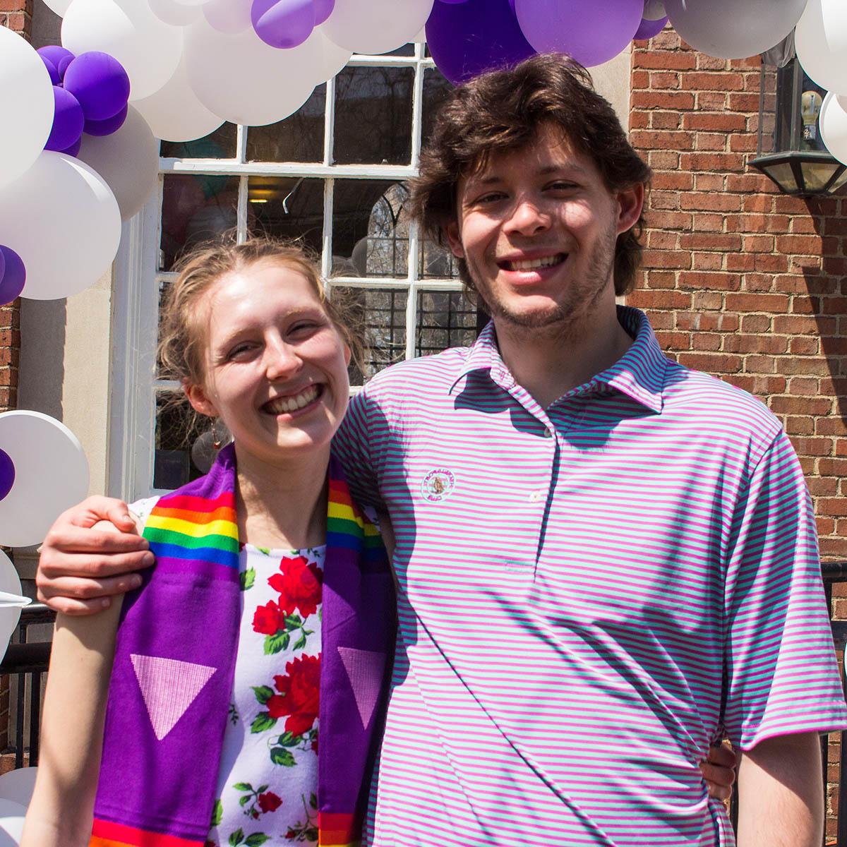 一名年轻女子穿着彩虹毕业礼服的照片, 与一名年轻男子合影并微笑，背景是紫色和白色的气球