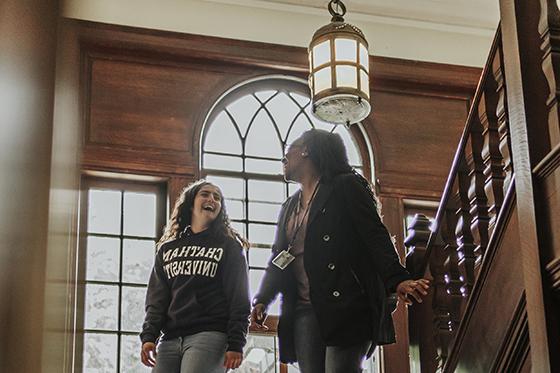 Photo of two female Chatham University students walk down wooden stairwell talking and laughing together. 