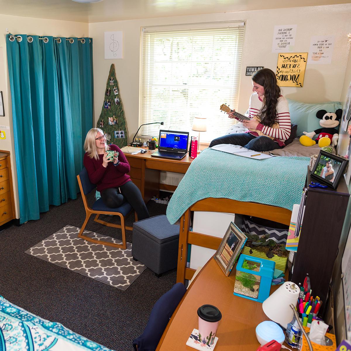 Photo of two roommates laughing and hanging out in their dorm room. One of them is playing a ukulele.
